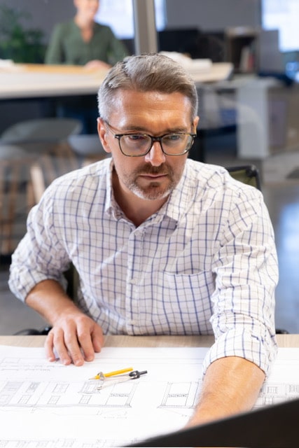 confident-caucasian-male-engineer-using-laptop-while-sitting-with-blueprint-desk-workplace